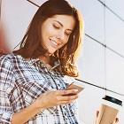 Eine Frau lehnt an der Wand und hat in einer Hand ein Smartphone, auf welches sie schaut, und in der anderen Hand ein To Go Becher.