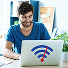 Ein junger Mann sitzt im Büro an seinem Schreibtisch. Vor ihm steht ein silbernen Laptop an dem er arbeitet. Auf dem Laptop ist ein WLAN-Zeichen in der Farbe blau und rot zu sehen.