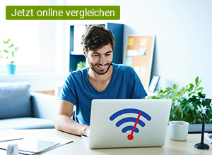 Ein junger Mann sitzt im Büro an seinem Schreibtisch. Vor ihm steht ein silbernen Laptop an dem er arbeitet. Auf dem Laptop ist ein WLAN-Zeichen in der Farbe blau und rot zu sehen.