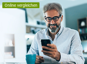 Ein Mann sitzt am Schreibtisch und schaut auf das Smartphone, welches sich in seiner Hand befindet, Auf dem Schreibtisch befindet sich ein Papierstapel, ein Kugelschreiber und eine Tasse.