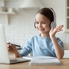 Ein junges Mädchen sitzt mit Kopfhörern an einem Tisch und hat einen Stift in der Hand. Vor ihr liegt ein weißer Block und es steht schräg vor ihr ein silbernen Laptop, auf welchen sie schaut und winkt.
