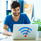 Ein junger Mann sitzt im Büro an seinem Schreibtisch. Vor ihm steht ein silbernen Laptop an dem er arbeitet. Auf dem Laptop ist ein WLAN-Zeichen in der Farbe blau und rot zu sehen.