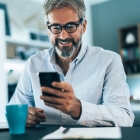 Ein Man sitzt am Schreibtisch und schaut auf das Smartphone, welches sich in seiner Hand befindet, Auf dem Schreibtisch befindet sich ein Papierstapel, ein Kugelschreiber und eine Tasse.