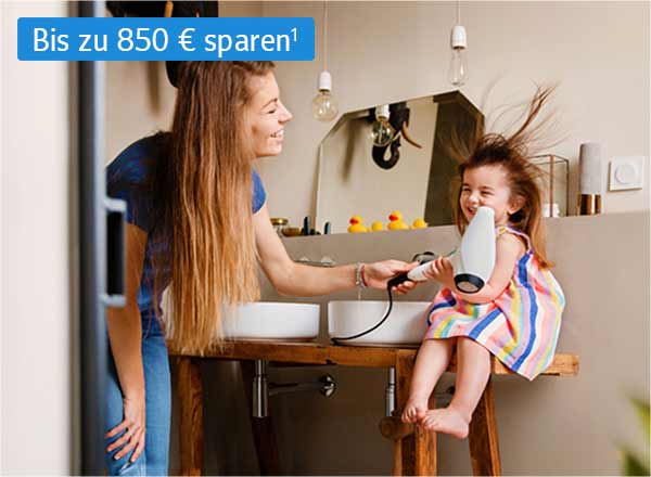 Eine Frau föhnt einem kleinen Mädchen in einem Badezimmer die Haare.  Die Haare des Mädchens sind durch den Fön zerzaust. Beide lächeln fröhlich.